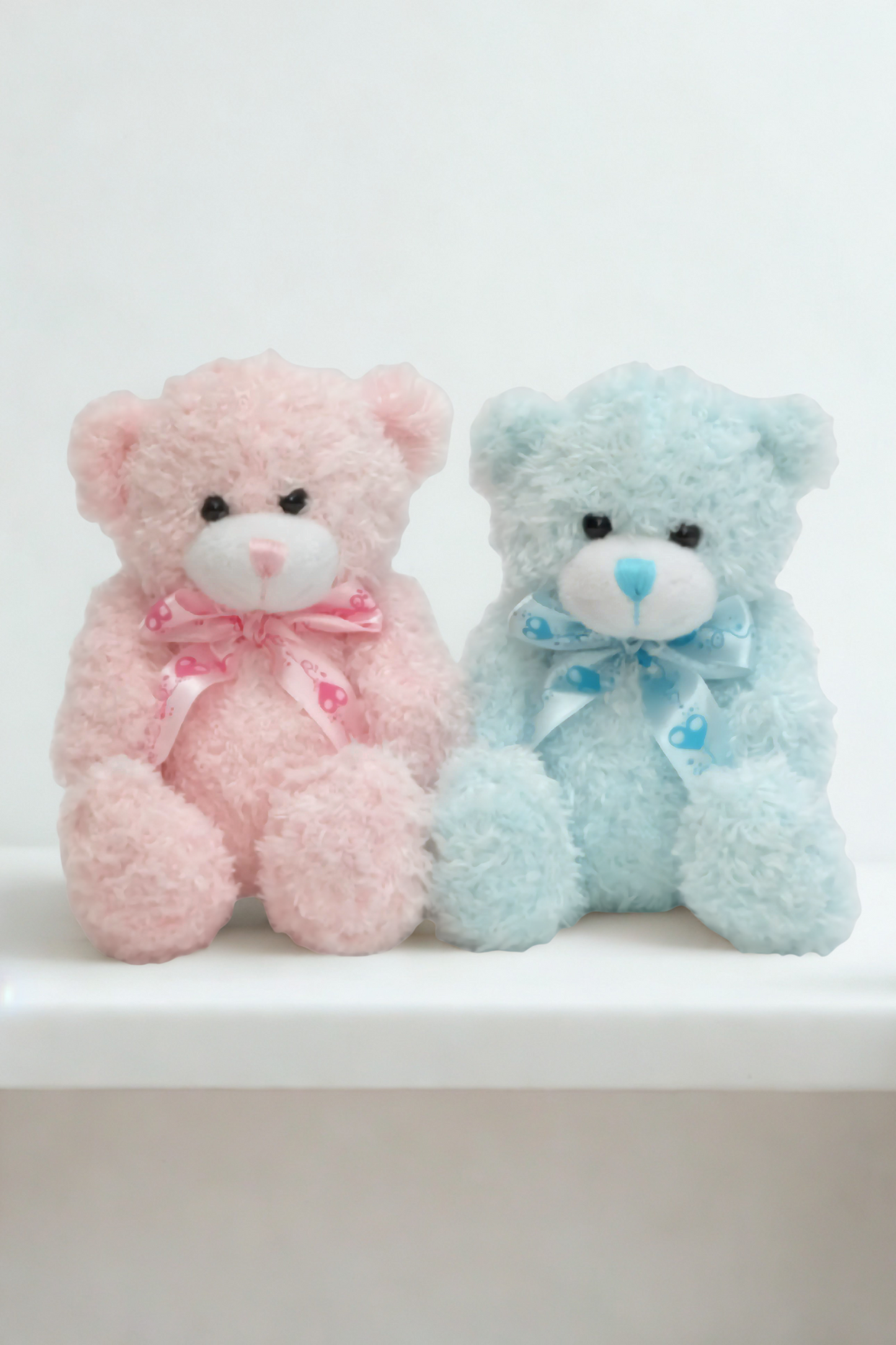 Two teddy bears, one pink and one blue, sitting side by side on a white surface with a white background.