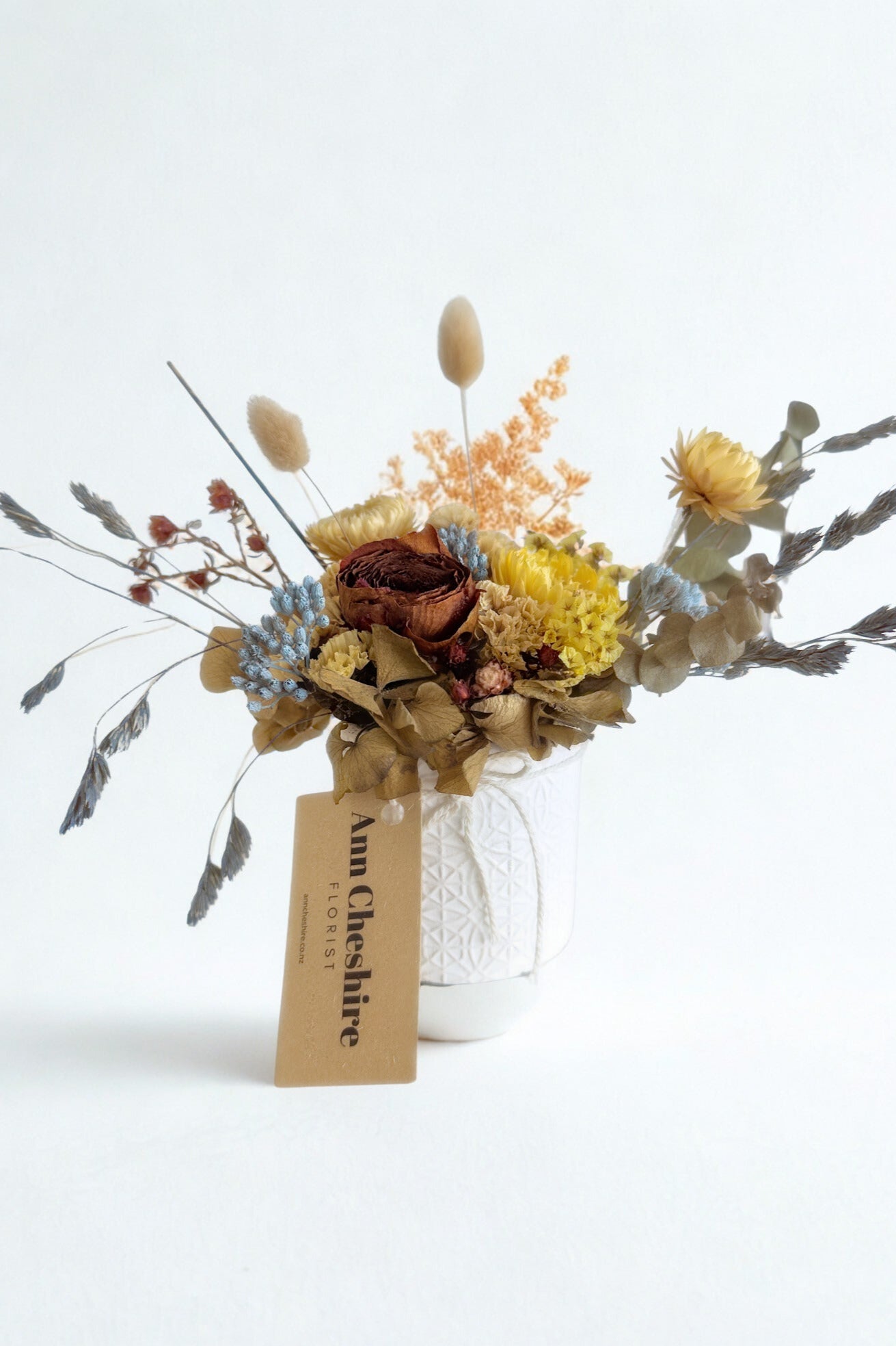 Dried flower arrangement in a glass vase with a visible brand tag on a white background
