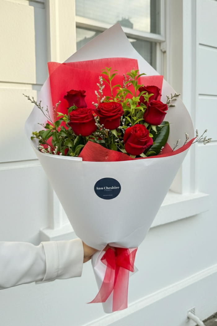 Bouquet of red roses wrapped in white paper with a red bow, held by a person against a white wall.
