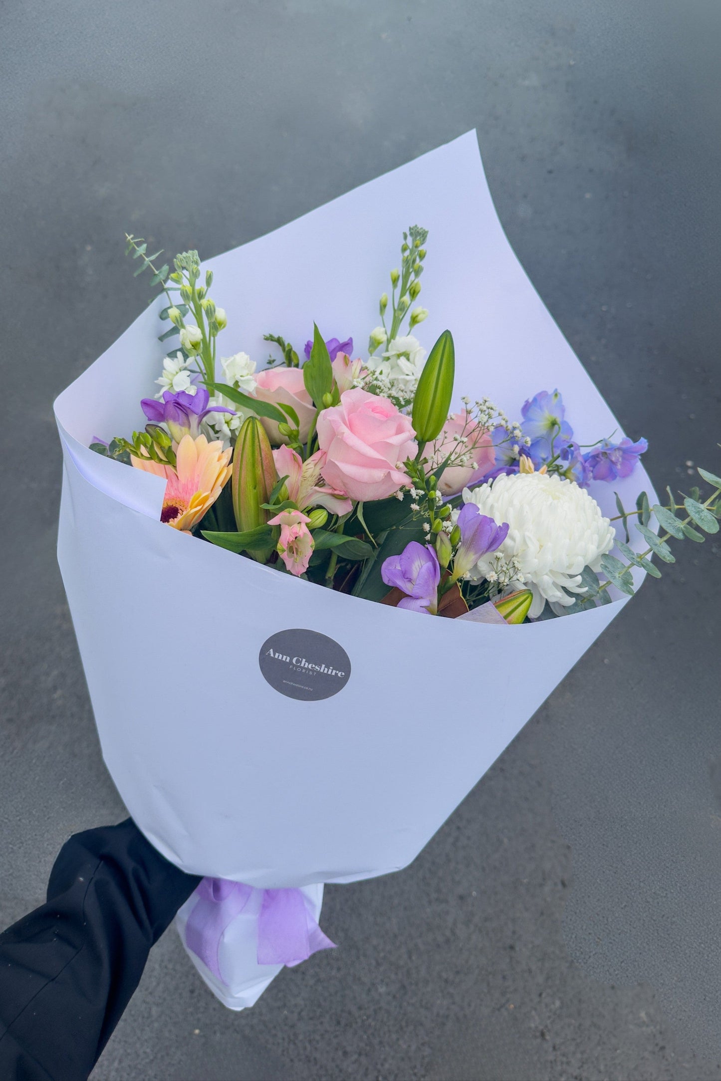 Bouquet of flowers wrapped in white paper with a brand label, held against a gray background.