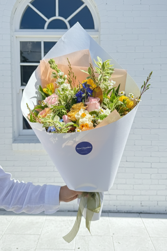 Bouquet of flowers held by a person in front of a white building with a blue window.