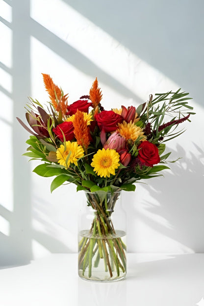 Bouquet of colorful flowers in a clear vase on a white surface with sunlight casting shadows.