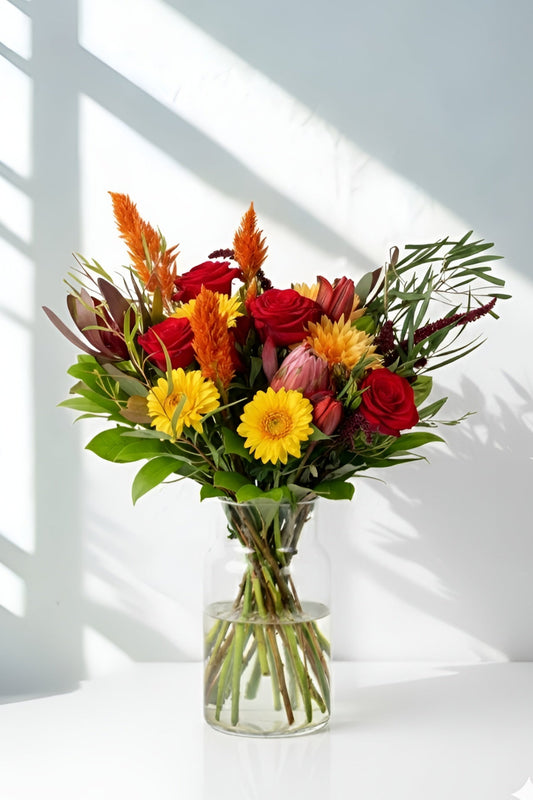 Bouquet of colorful flowers in a clear vase on a white surface with sunlight casting shadows.
