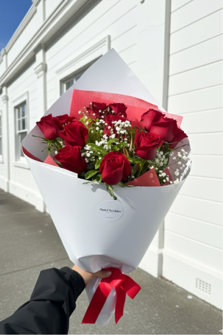 Bouquet of red roses with a white wrap and red ribbon held against a white building.