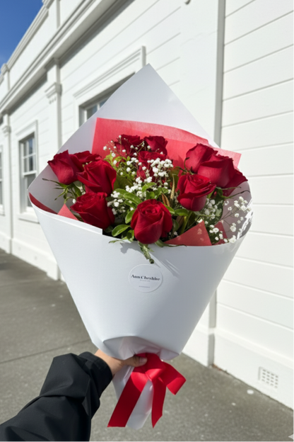 Bouquet of red roses with a white wrap and red ribbon held against a white building.