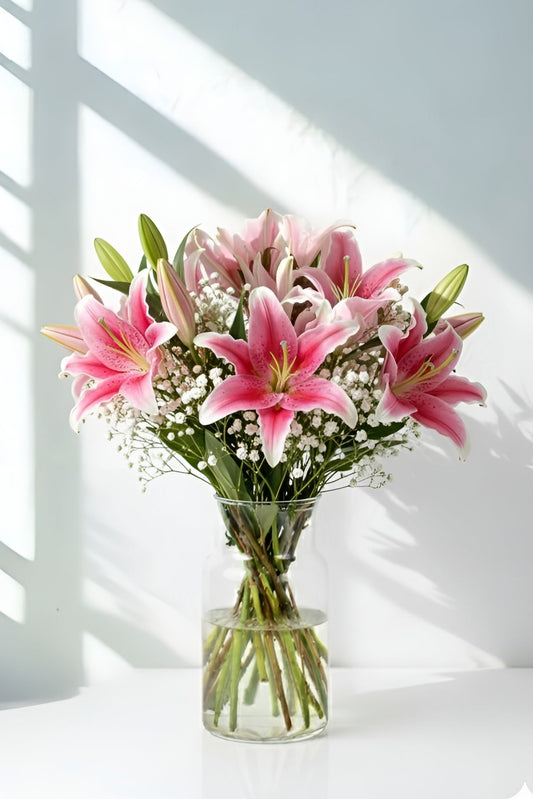 Bouquet of pink lilies in a clear vase on a white surface with sunlight casting shadows.