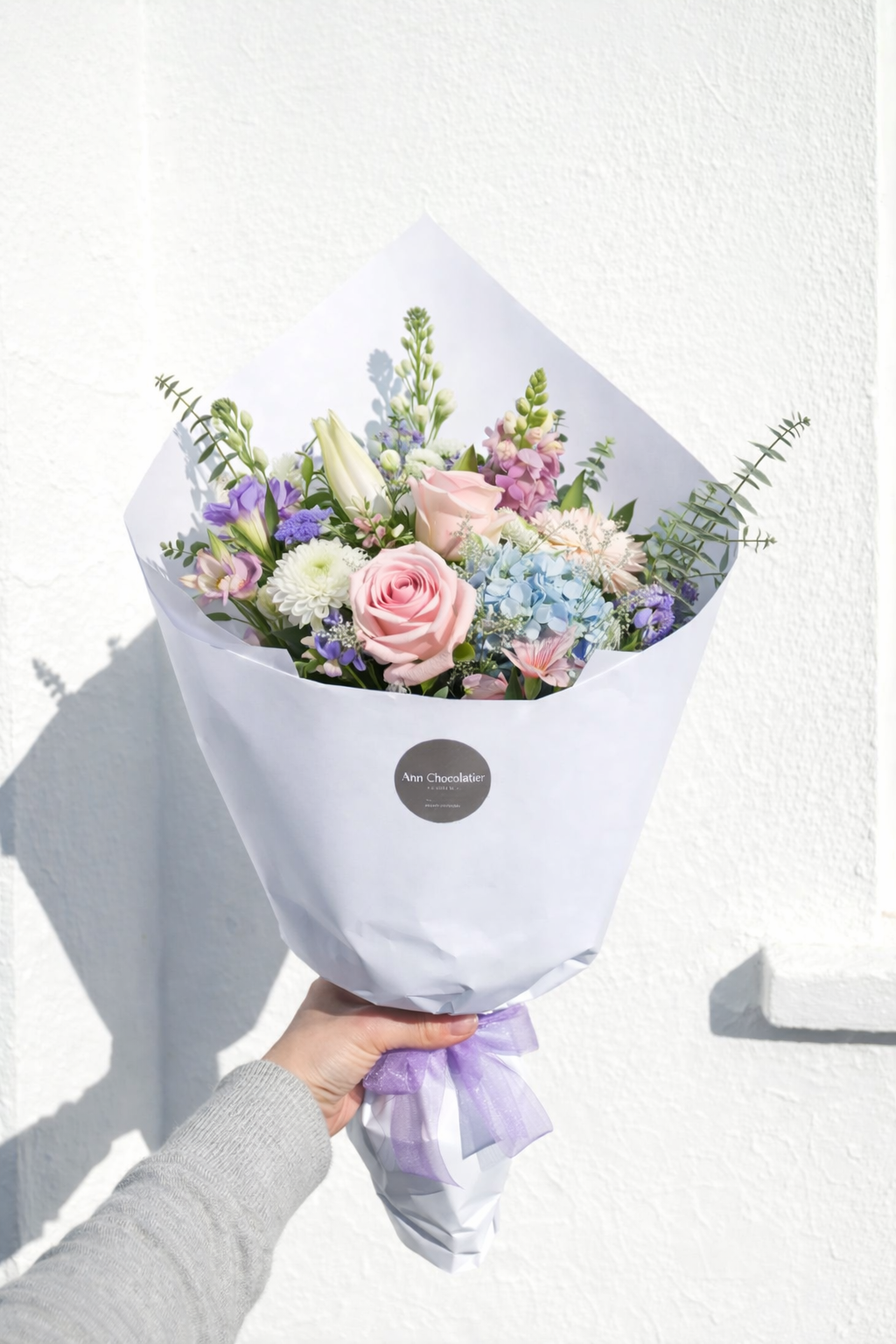 Bouquet of flowers wrapped in white paper with a purple ribbon, held against a white background.