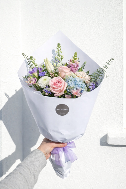 Bouquet of flowers wrapped in white paper with a purple ribbon, held against a white background.