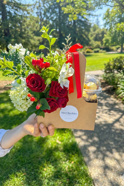 Bouquet of red roses and greenery in a brown paper bag with a red ribbon, held outdoors.