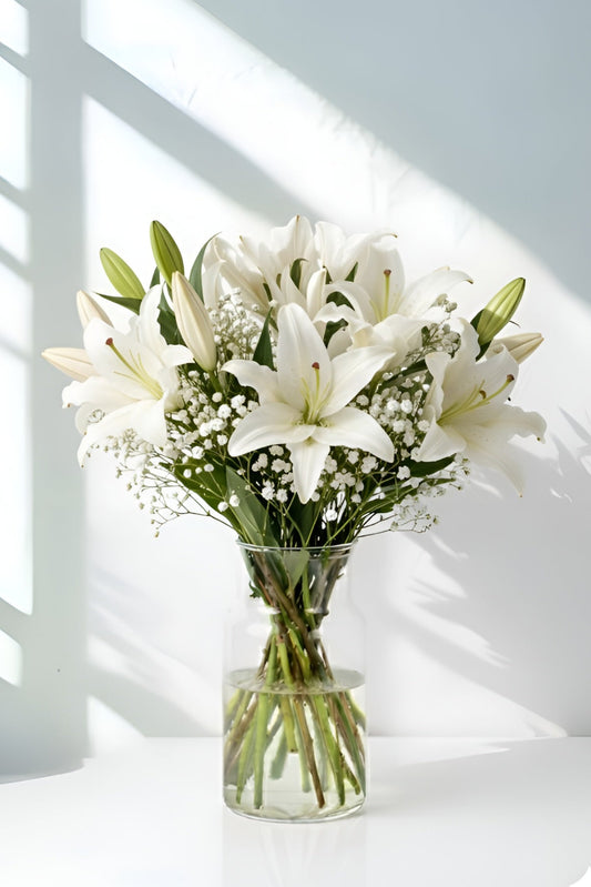 Bouquet of white flowers in a clear vase on a light surface with shadows from a window.