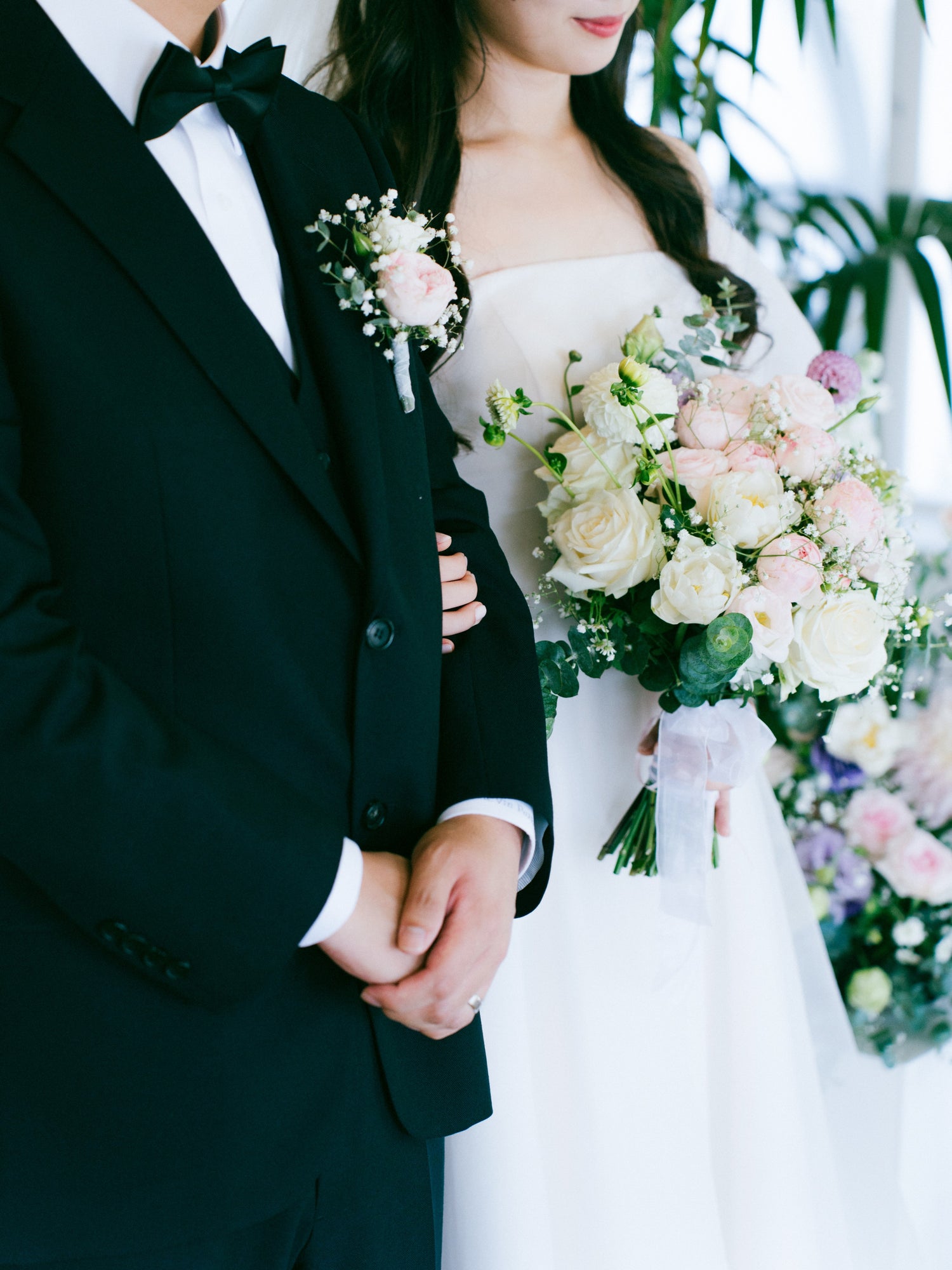 A bride and groom standing together, the bride holding a pastel-toned bouquet of roses and greenery, and the groom wearing a black tuxedo with a matching boutonniere.