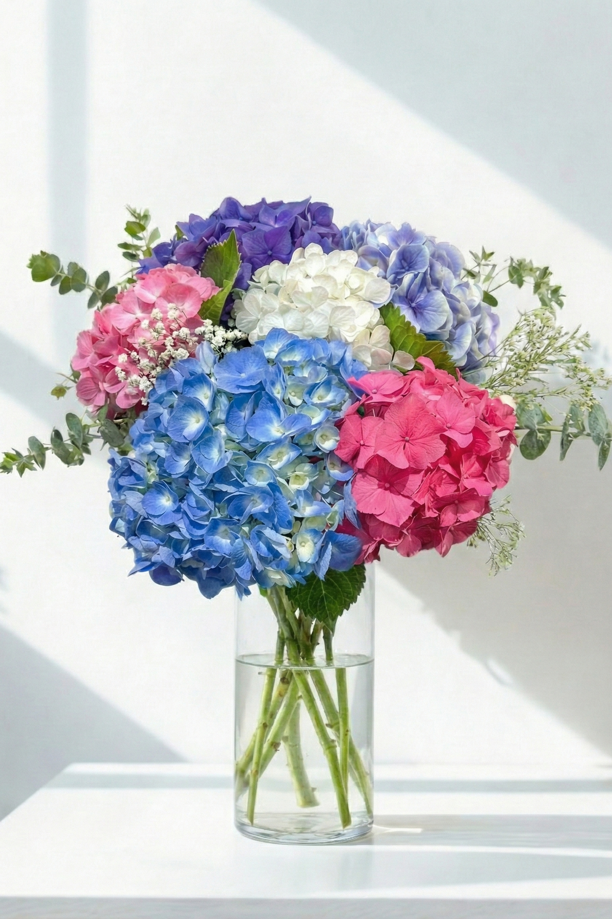Bouquet of pink, blue, and white hydrangeas in a clear vase on a white surface.